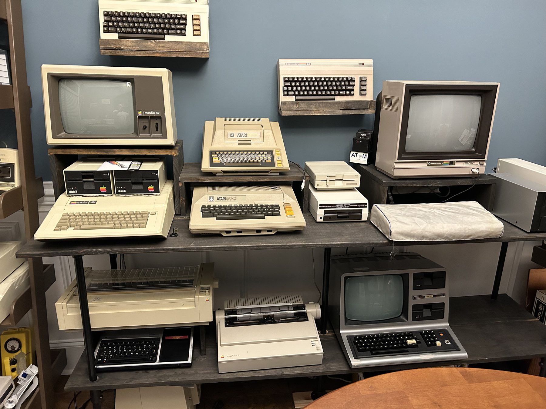 A shelf of vintage computers: a Commodore 64, Atari 400 and 800, Apple IIe with two Disk II drives and a Monitor III, a Commodore 1541 disk drive and monitor, a TRS-80 Model III, a TI-99/4A, and Apple dot matrix printers.